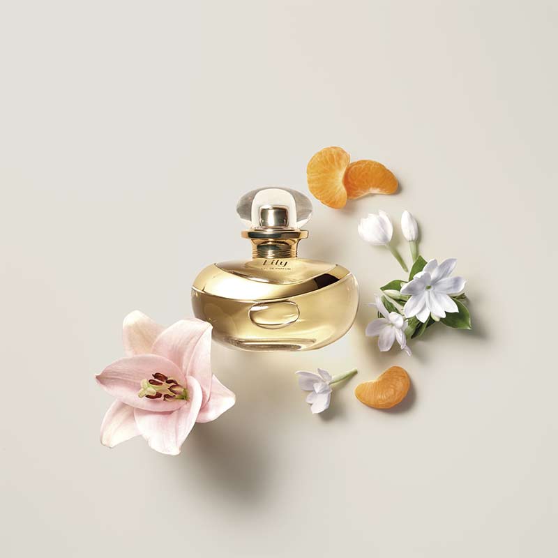 Frasco do perfume Lily 75ml do Boticário, dourado e arredondado, rodeado por lírio rosa, flores brancas e gomos de tangerina sobre fundo claro.