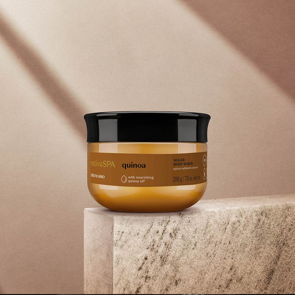 Açúcar Esfoliante Nativa SPA Quinoa – oBoticário, pote dourado com tampa preta sobre pedra em fundo bege, iluminado por luz suave, representando cuidado e nutrição corporal.