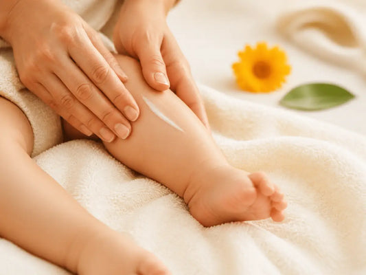 Mãos de um adulto aplicam loção hidratante na perna de um bebé, sobre uma toalha suave, com calêndula ao fundo.
