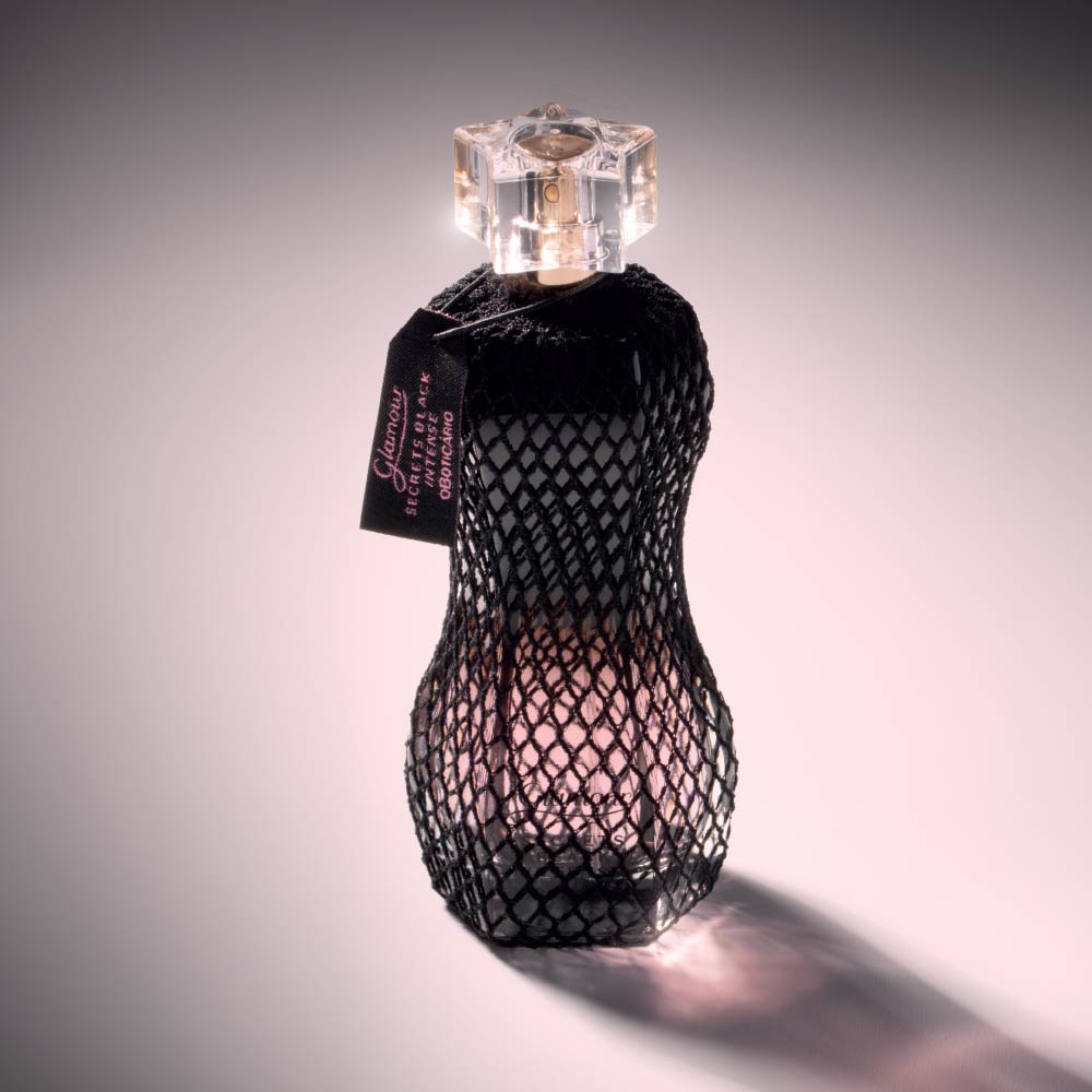 Perfume Glamour Secrets Black Intense 75ml do Boticário, com frasco em formato curvo protegido por rede preta, iluminado em fundo suave.