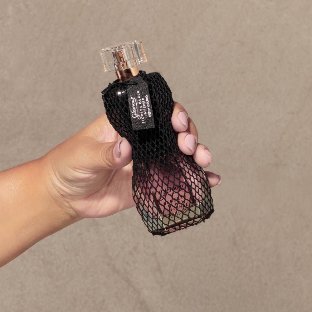Mão a segurar o perfume Glamour Secrets Black Intense 75ml do Boticário, mostrando o frasco em vidro com rede preta.