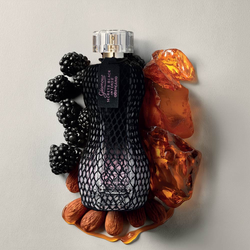 Frasco do perfume Glamour Secrets Black Intense 75ml do Boticário, envolvido em rede preta, colocado entre amoras, pedras de âmbar e amêndoas.