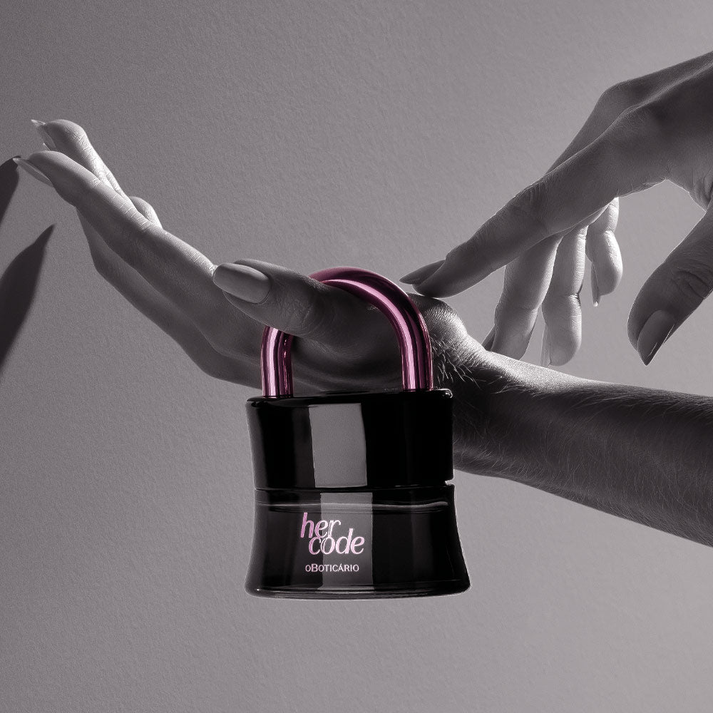 Perfume Her Code Touch 50ml do Boticário, frasco em forma de cadeado com arco rosa metálico, segurado entre duas mãos em fundo cinza.