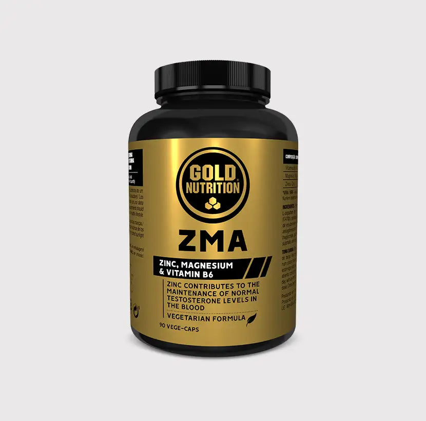 Frasco preto e dourado de ZMA GoldNutrition, com 90 cápsulas, fotografado num ginásio junto a halteres.